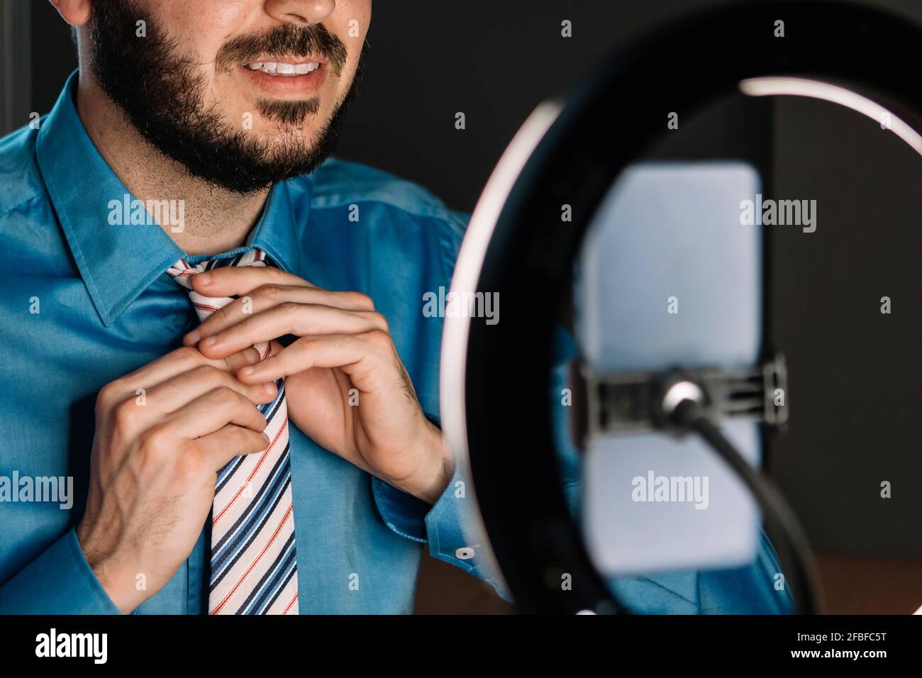 Mid adult influencer adjusting necktie while vlogging through mobile phone with illuminated camera flash at studio Stock Photo