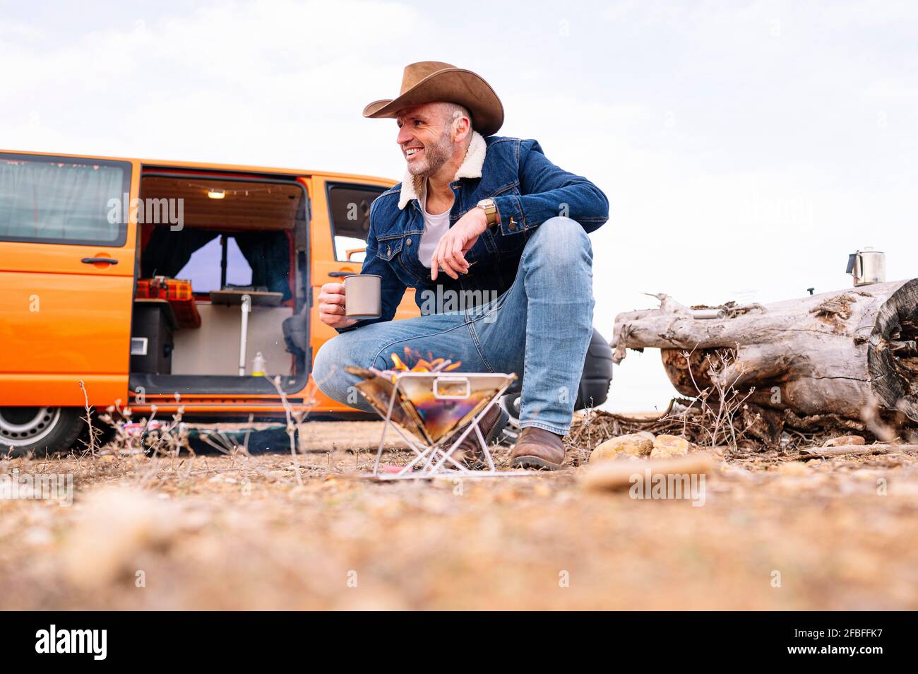 https://c8.alamy.com/comp/2FBFFK7/smiling-mature-man-in-hat-crouching-while-having-coffee-by-wood-burning-stove-during-camp-2FBFFK7.jpg