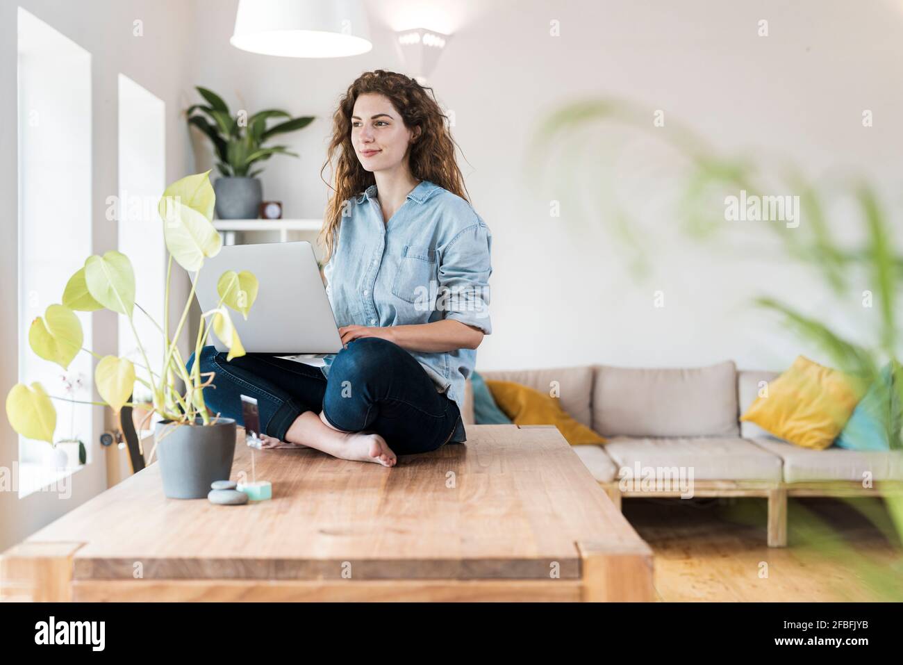 Contented woman with laptop day dreaming while sitting on table at home Stock Photo