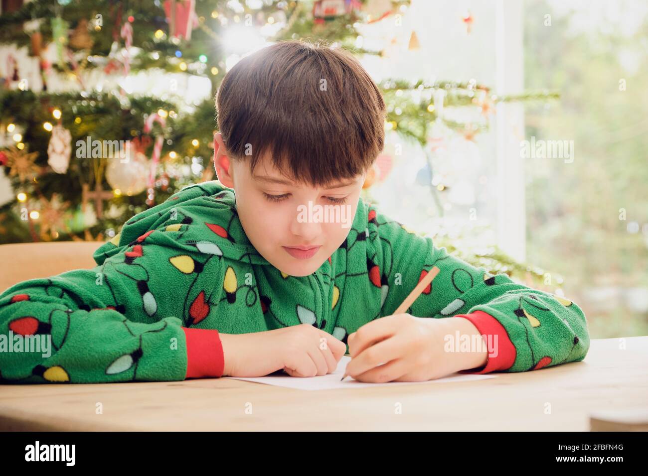 Cute boy writing on paper at table with Christmas tree in background at home Stock Photo