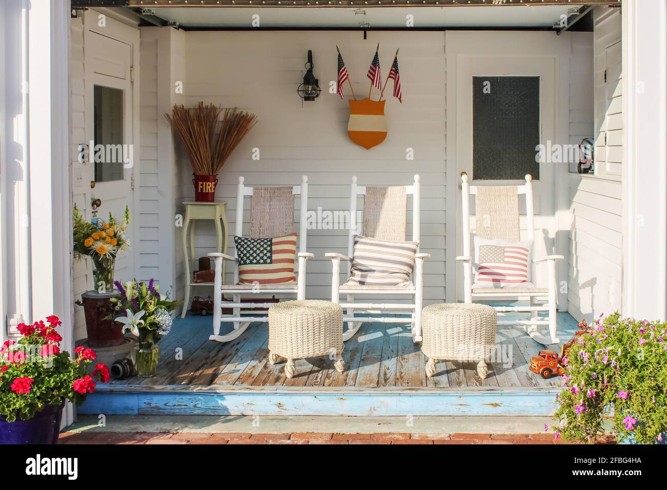 Fireman and patriotic themed worn wooden front porch in Cape Cod with three white rocking chairs and pillows and wicker foot stools and flags and pots Stock Photo