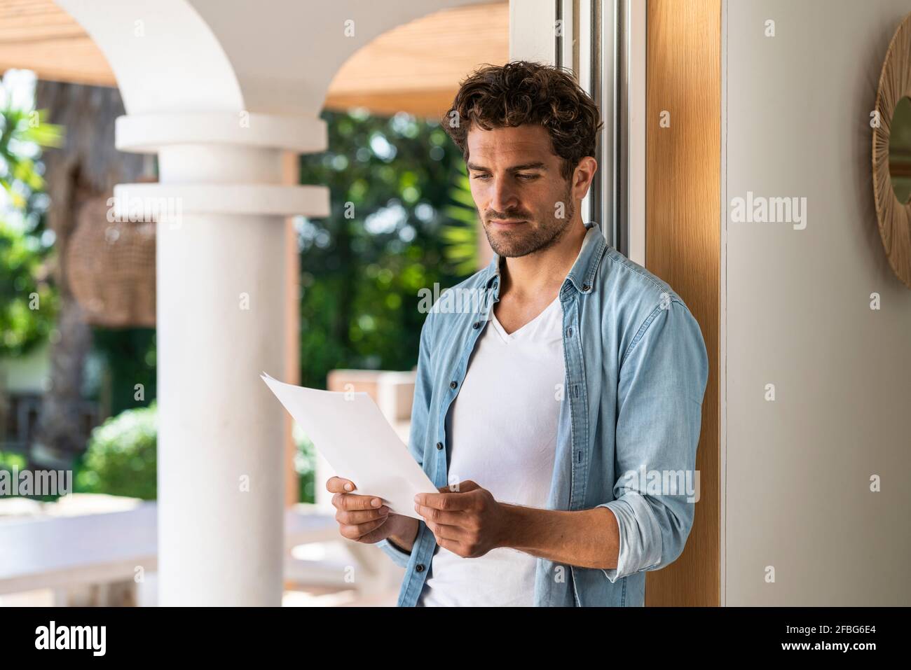 Serious man reading document while leaning on doorframe at home Stock Photo