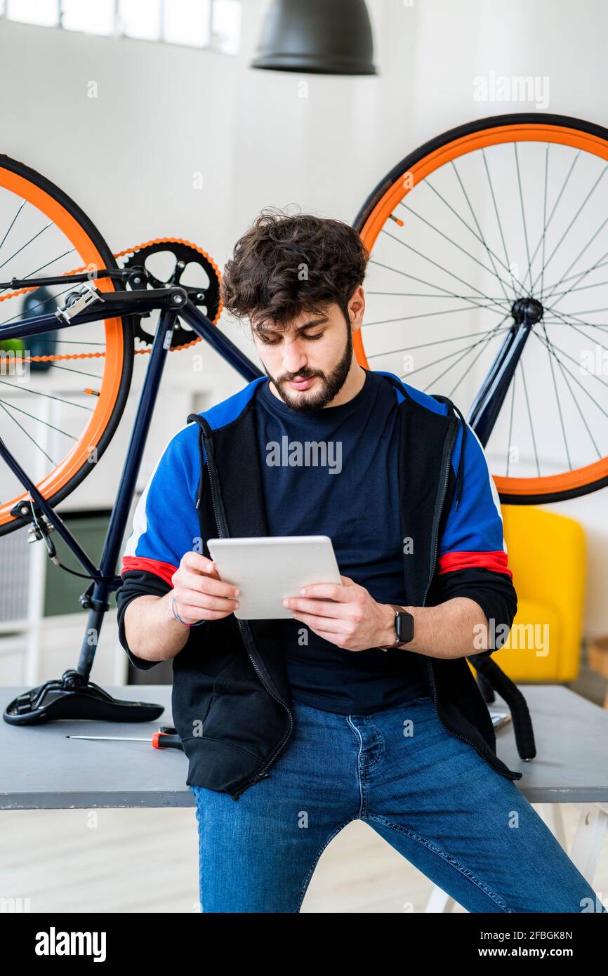 Man using digital tablet while leaning on table with upturnt bicycle at home Stock Photo
