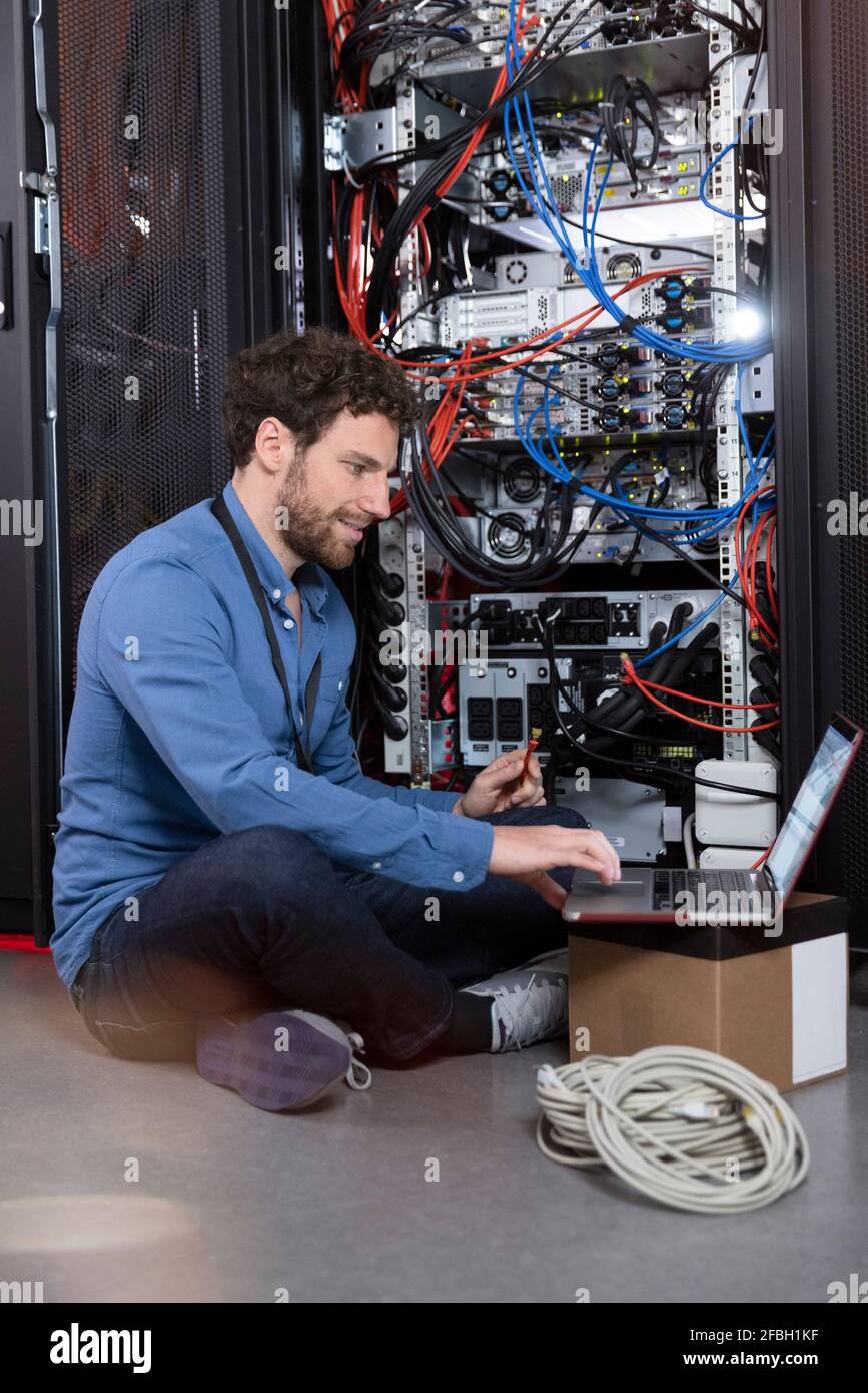 Smiling male IT specialist holding patch cord cable while using laptop in data center Stock Photo