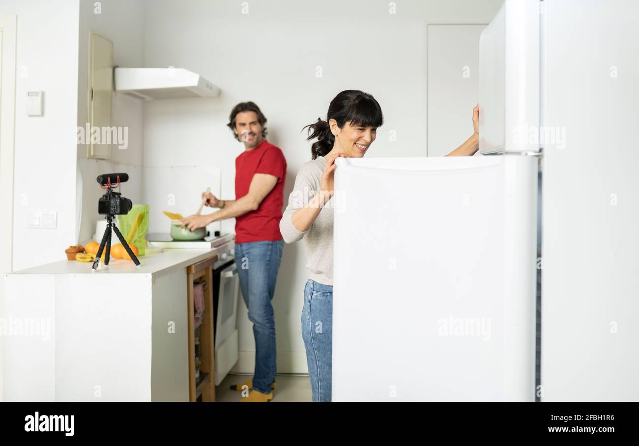 Smiling woman opening refrigerator door while man preparing food in kitchen at home Stock Photo