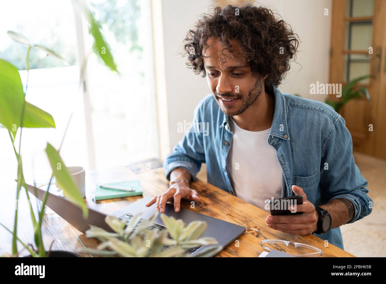 Young businessman holding smart phone while using laptop at home Stock Photo
