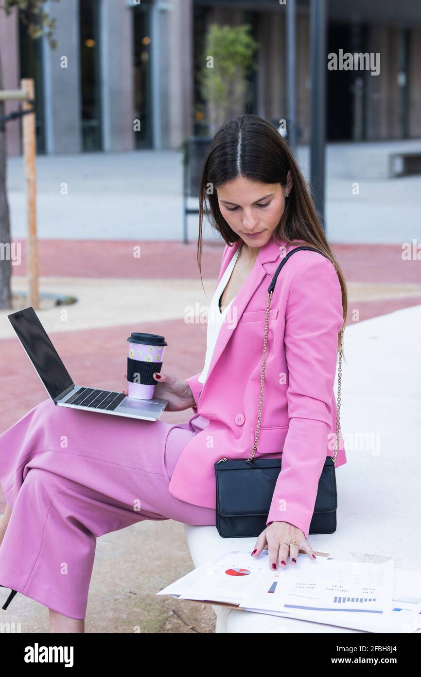Young female entrepreneur with laptop checking papers while sitting on retaining wall Stock Photo