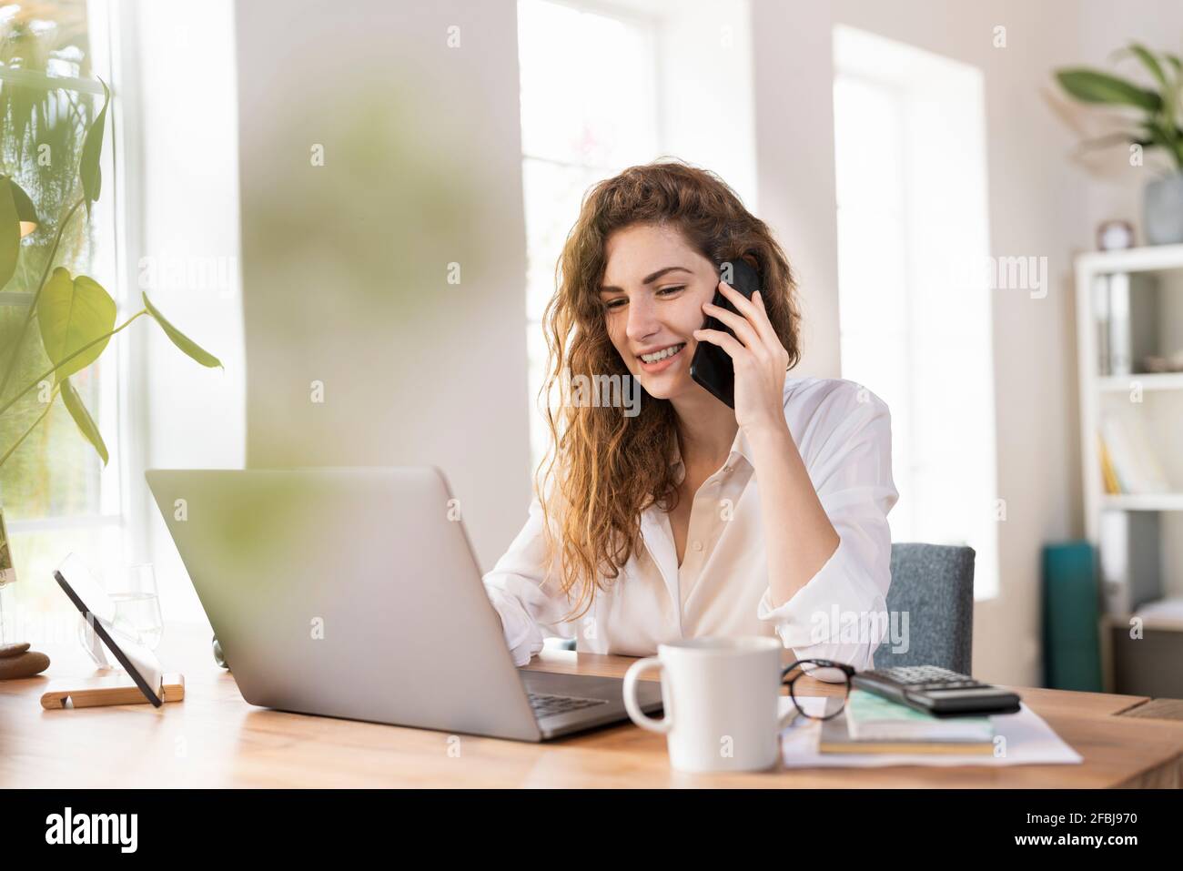 Contented female entrepreneur talking on smart phone while using laptop at desk in home office Stock Photo