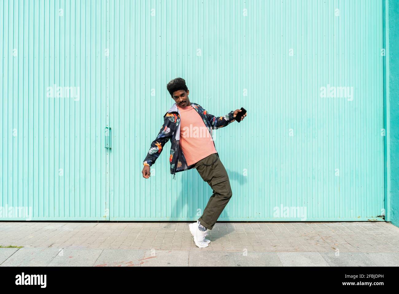 Ecstatic man holding smart phone while dancing in front of turquoise wall Stock Photo