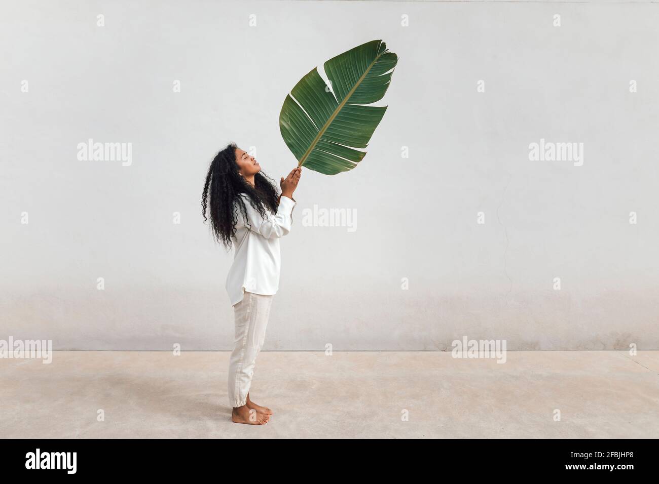 Young woman with curly hair holding big green leaf standing by white wall Stock Photo