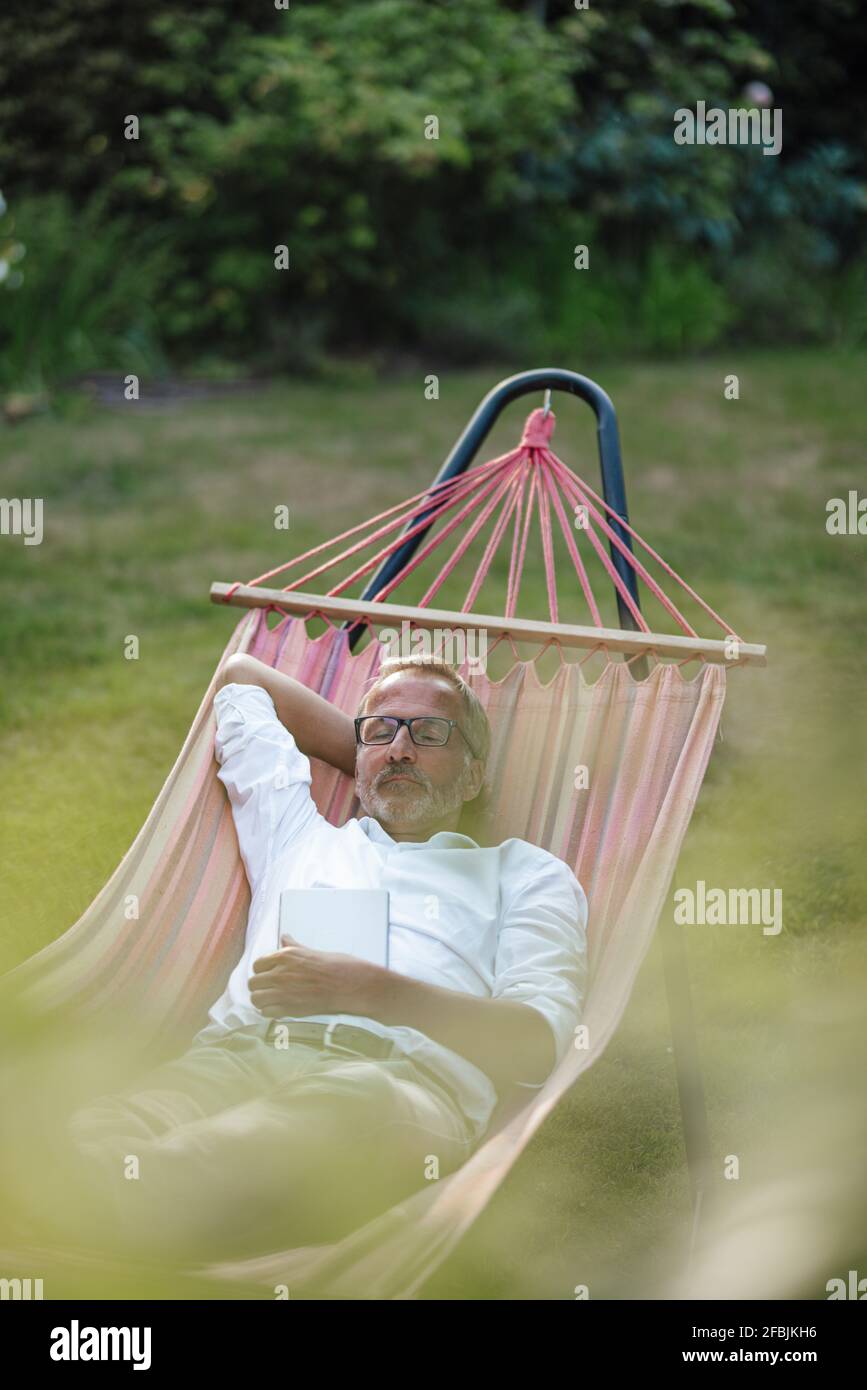 Mature man with book relaxing on hammock in backyard Stock Photo