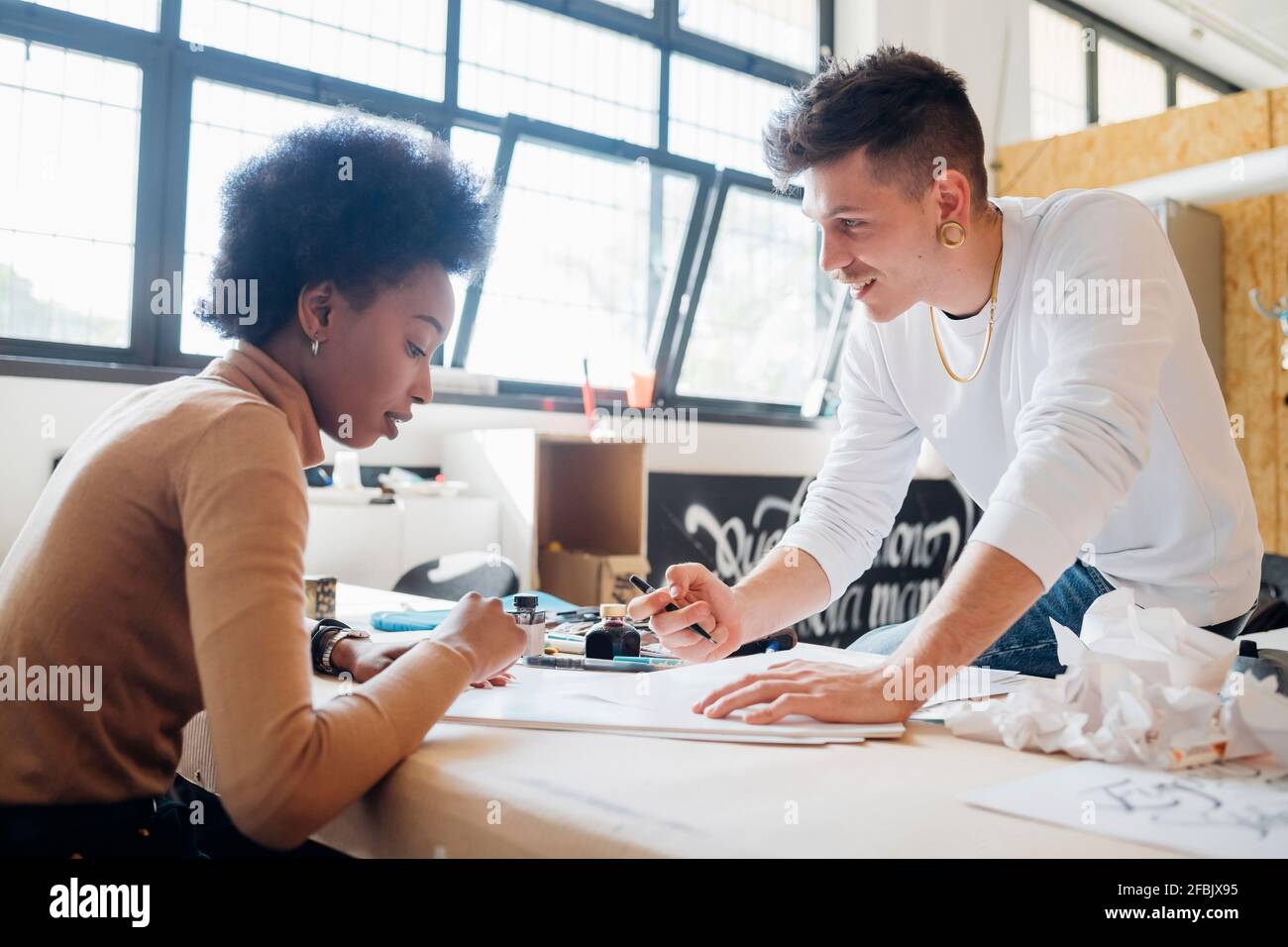 Smiling hipster man teaching girlfriend calligraphy while sitting at table in studio Stock Photo