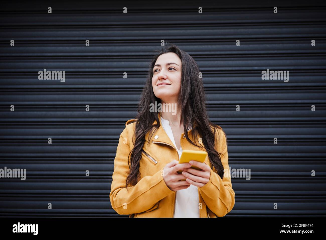 https://c8.alamy.com/comp/2FBK474/smiling-woman-with-mobile-phone-looking-away-while-standing-in-front-of-black-shutter-2FBK474.jpg