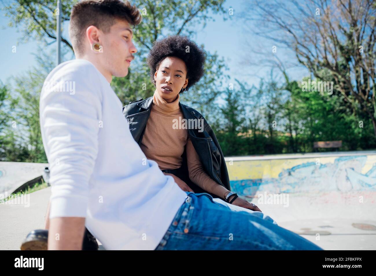 Multi ethnic couple sitting at skateboard park during sunny day Stock Photo