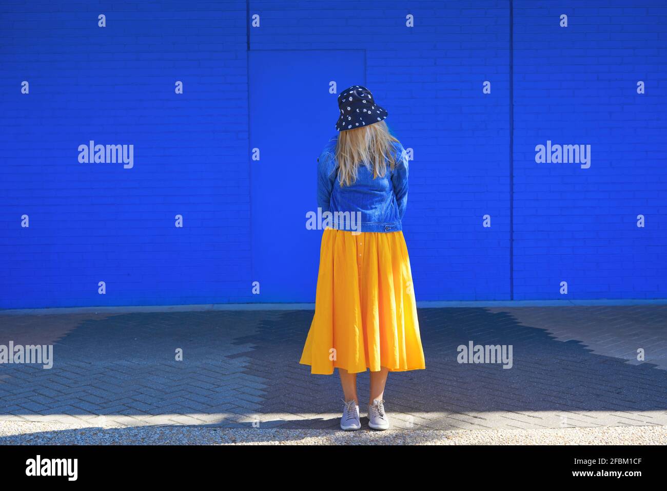 Caucasian woman wearing bucket hat and denim jacket while standing in front of blue wall Stock Photo