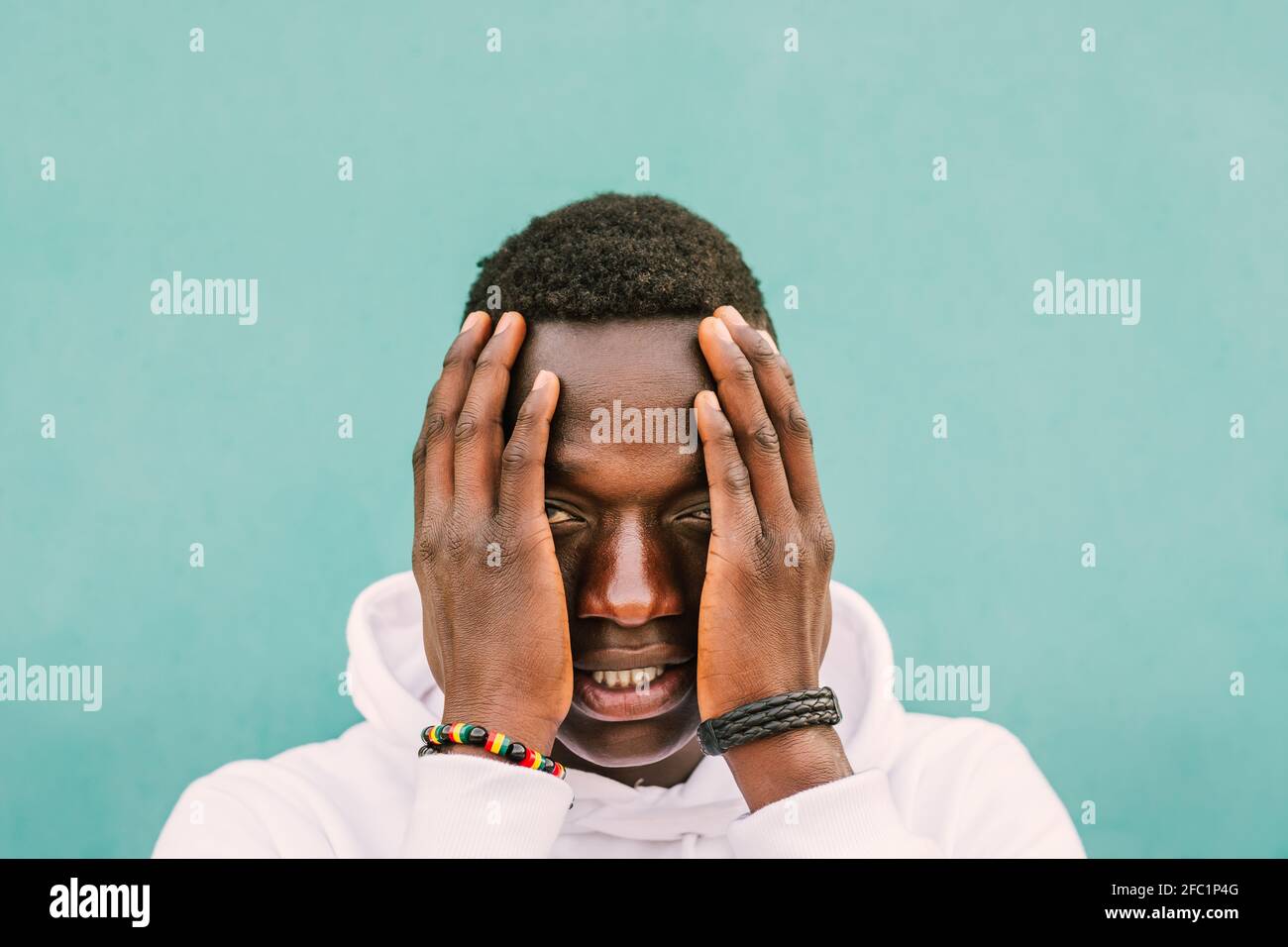 Portrait of young african black man against a green wall with white sweatshirt with his hands on his face. Stop racism protest concept. BLM Stock Photo