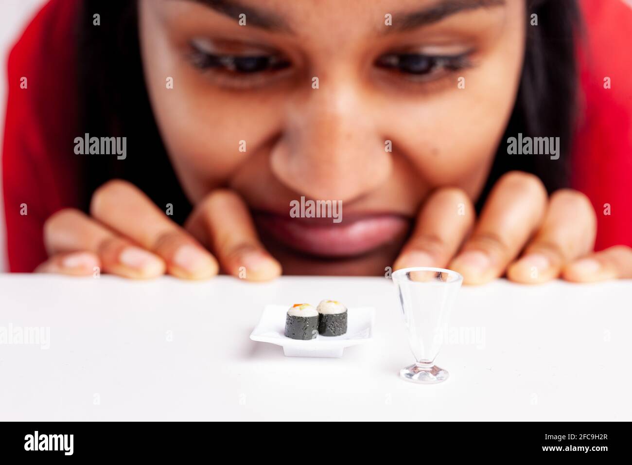 Young woman with an aversion to sushi in a fun concept using miniature food on a plate with her peering over the table grimacing and selective focus t Stock Photo
