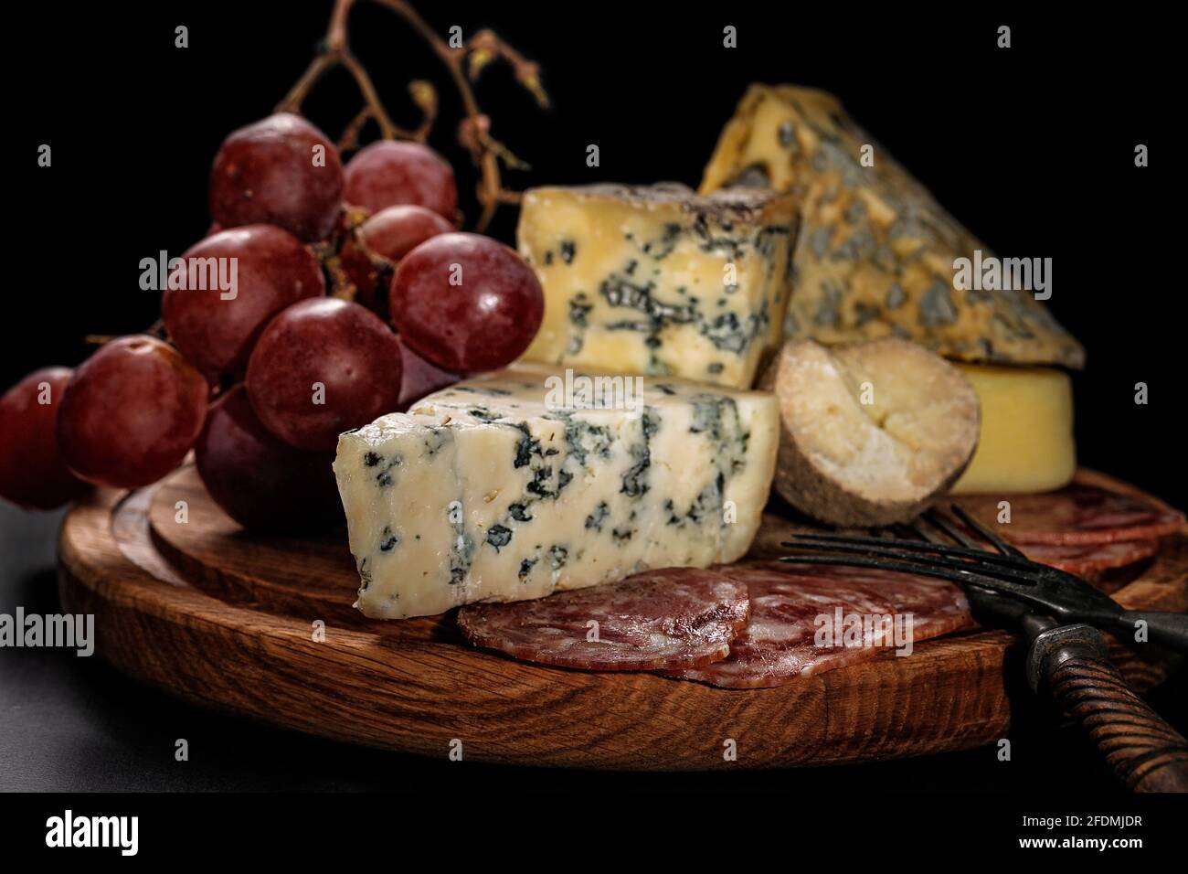 Food still life. Assorted various gourmet cheeses, raw smoked sausages and large sweet grapes on a dark background. Various types of cheese: hard and Stock Photo