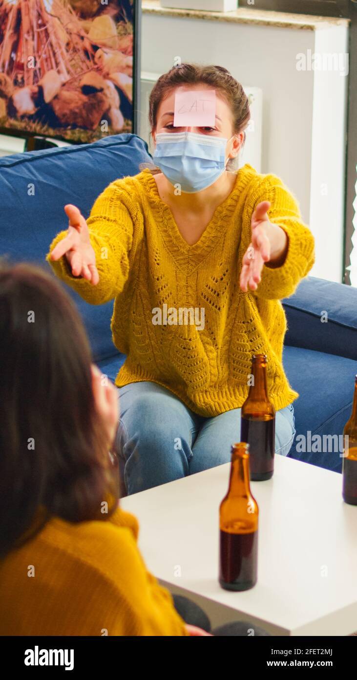 Funny friends with sticky notes on foreheads enjoying name game gesturing  during new normal party wearing face mask keeping social distancing  preventing covid19 spread. People enjoying bottles of beer Stock Photo -