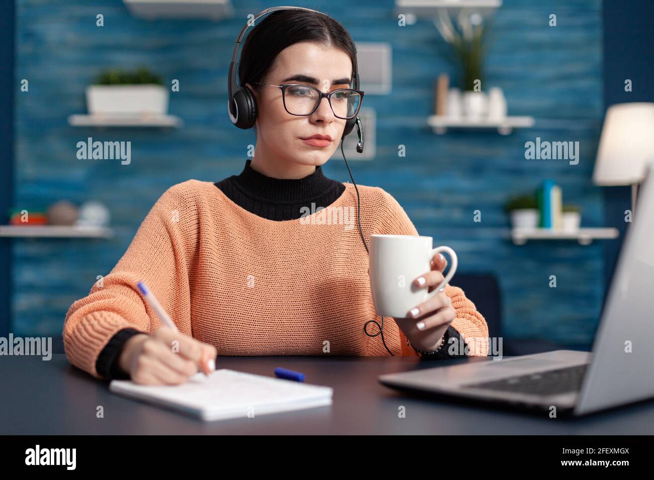 College student taking notes on paper while looking at university website searching information for online course. Youg woman using laptop computer while sitting at desk in living room Stock Photo