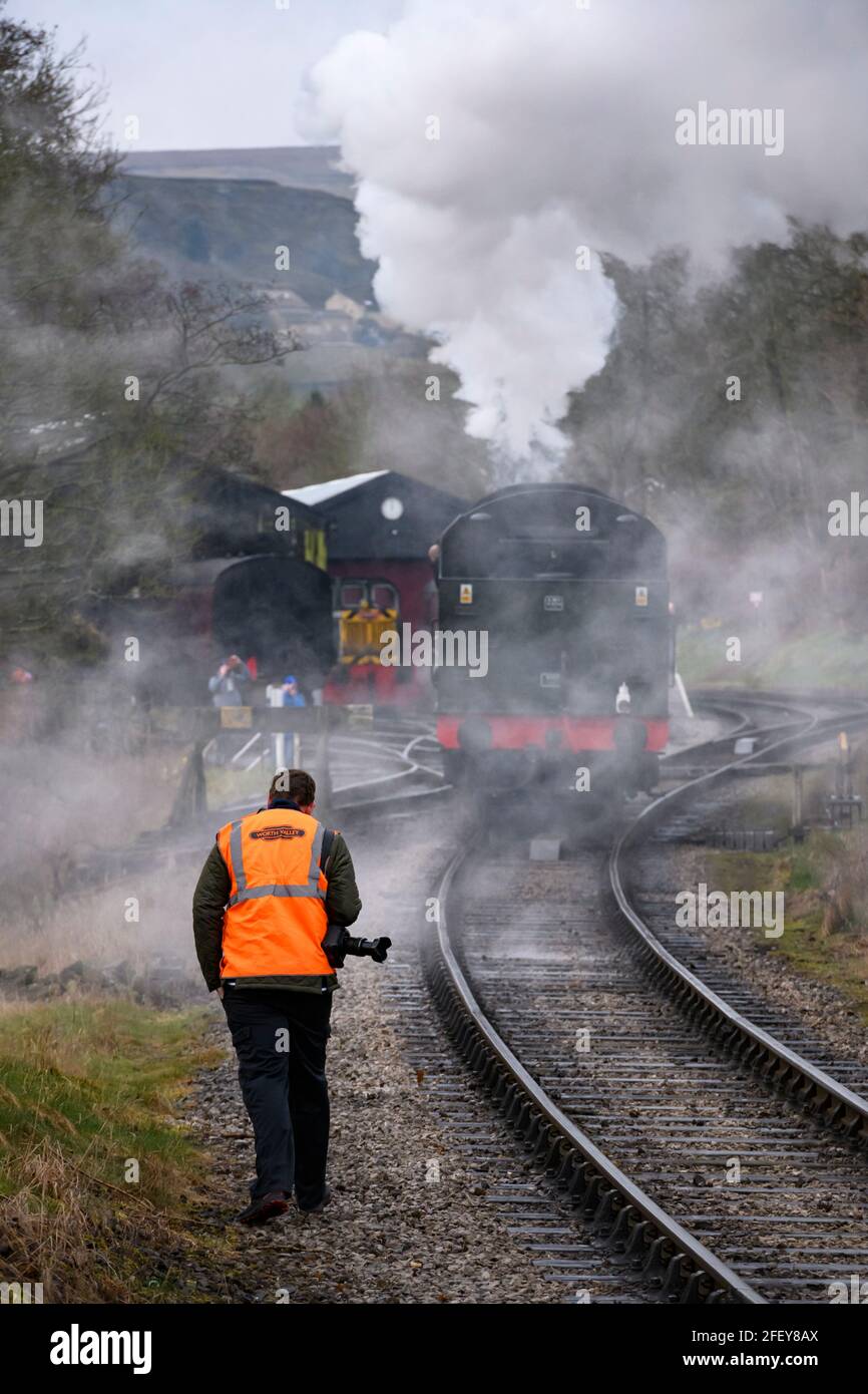 Historic steam train or loco puffing dramatic clouds of smoke (trackside enthusiasts with cameras) - Oxenhope Station sidings, Yorkshire, England, UK. Stock Photo