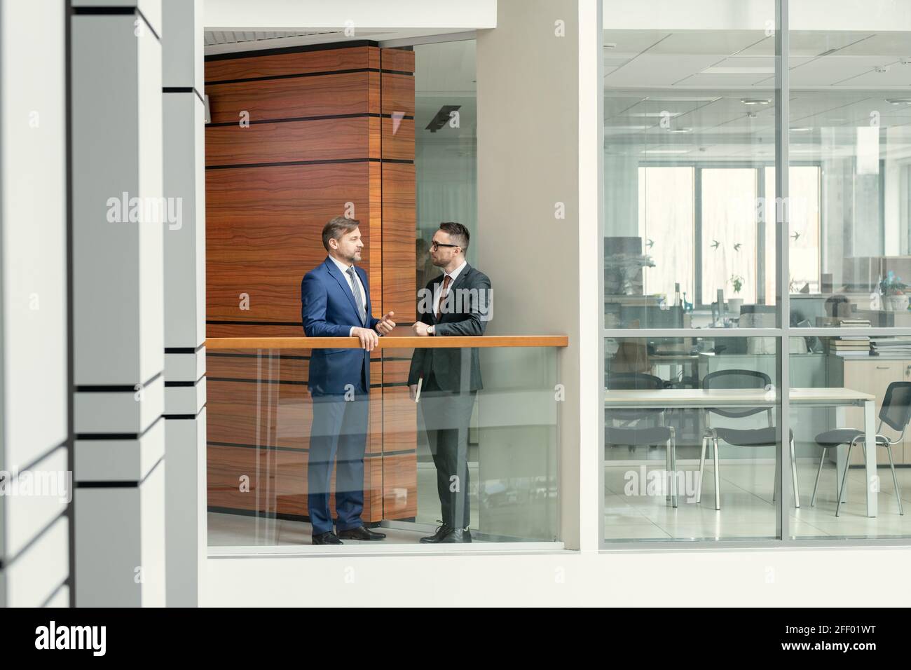Mature businessman standing at railing and talking to young colleague in corridor Stock Photo