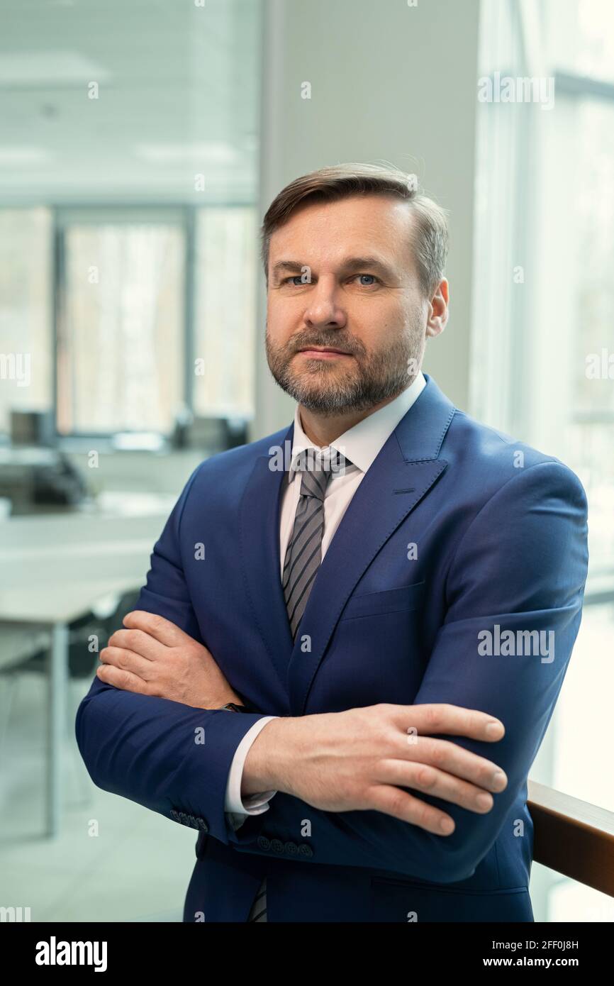 Portrait of content bearded manager in dark blue suit standing with crossed arms confidently against office room Stock Photo