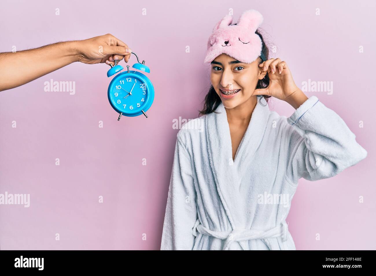 Hispanic teenager girl with dental braces wearing sleep mask and robe with  alarm clock close to her smiling with hand over ear listening and hearing t  Stock Photo - Alamy