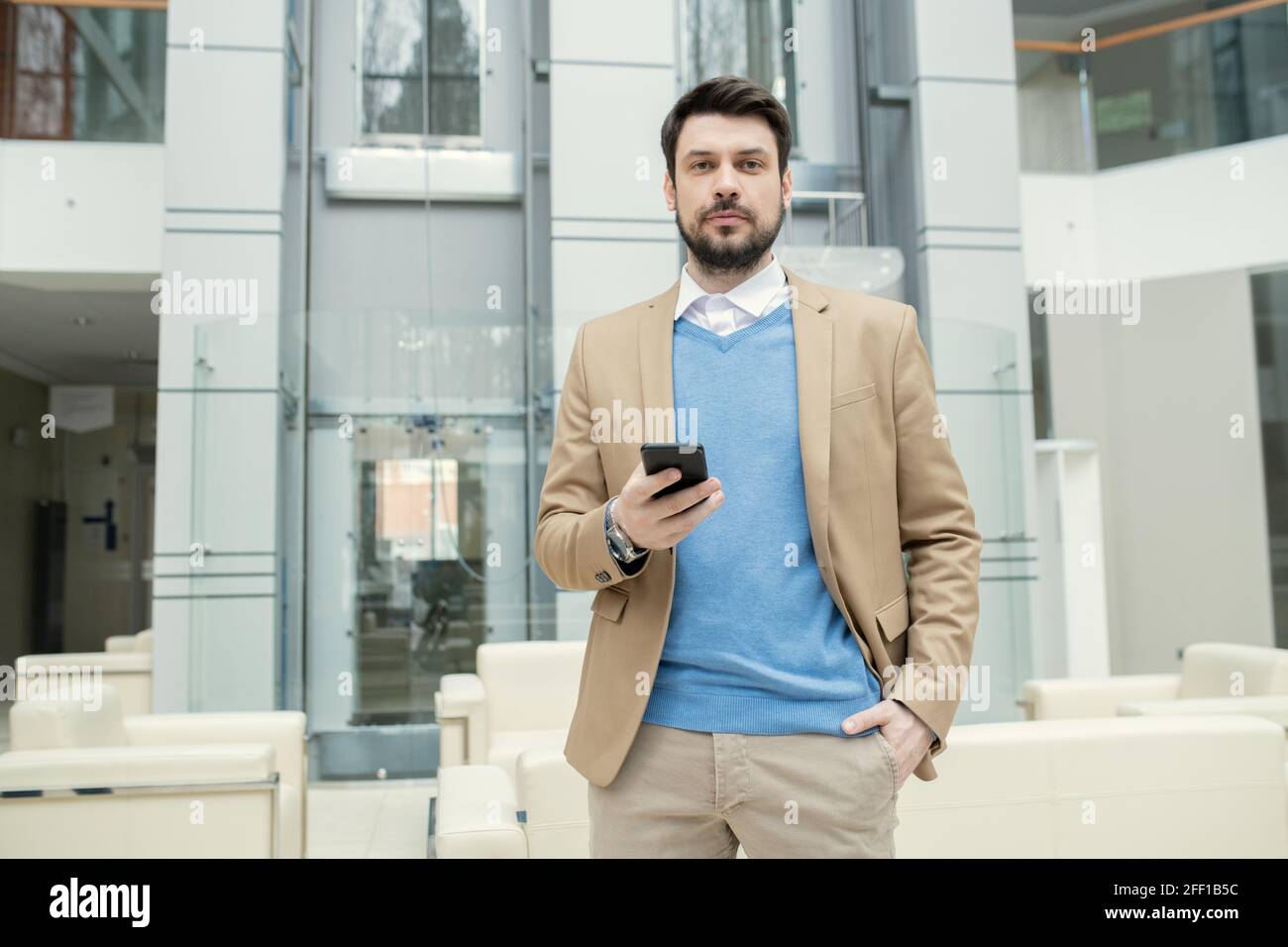 Portrait of content young bearded lawyer in blue sweater holding hand in pocket and checking smartphone in lobby Stock Photo
