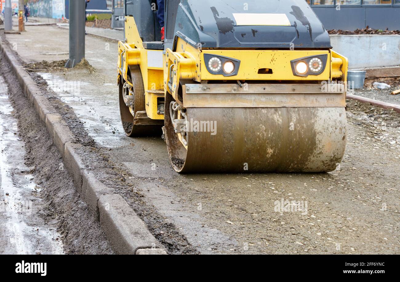 Frontal view of a road roller rolling the base of the sidewalk along a concrete curb near the carriageway. Stock Photo