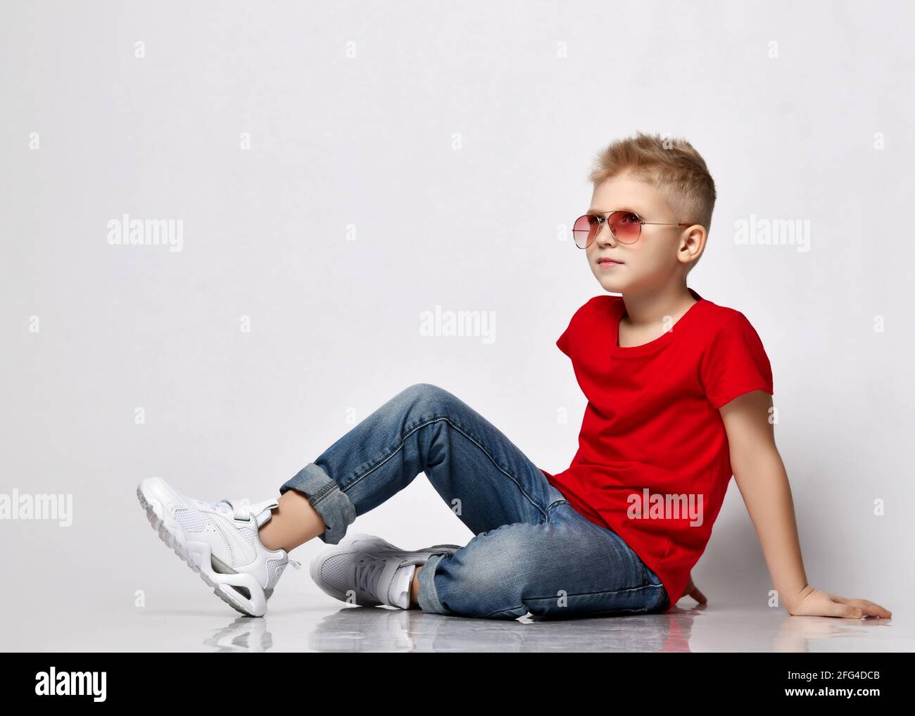Regular blond kid boy in red t-shirt, blue jeans, white sneakers and sunglasses sits on floor leaning on his hands Stock Photo