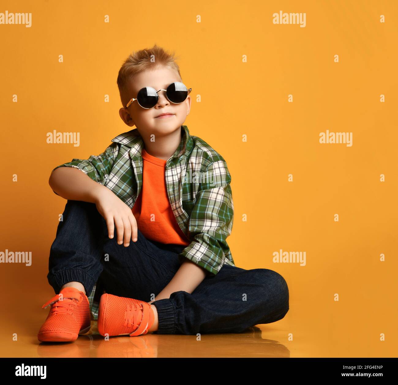 Calm nice kid, schoolboy teenager in round sunglasses, checkered shirt, t-shirt and jeans sits on floor looking up Stock Photo