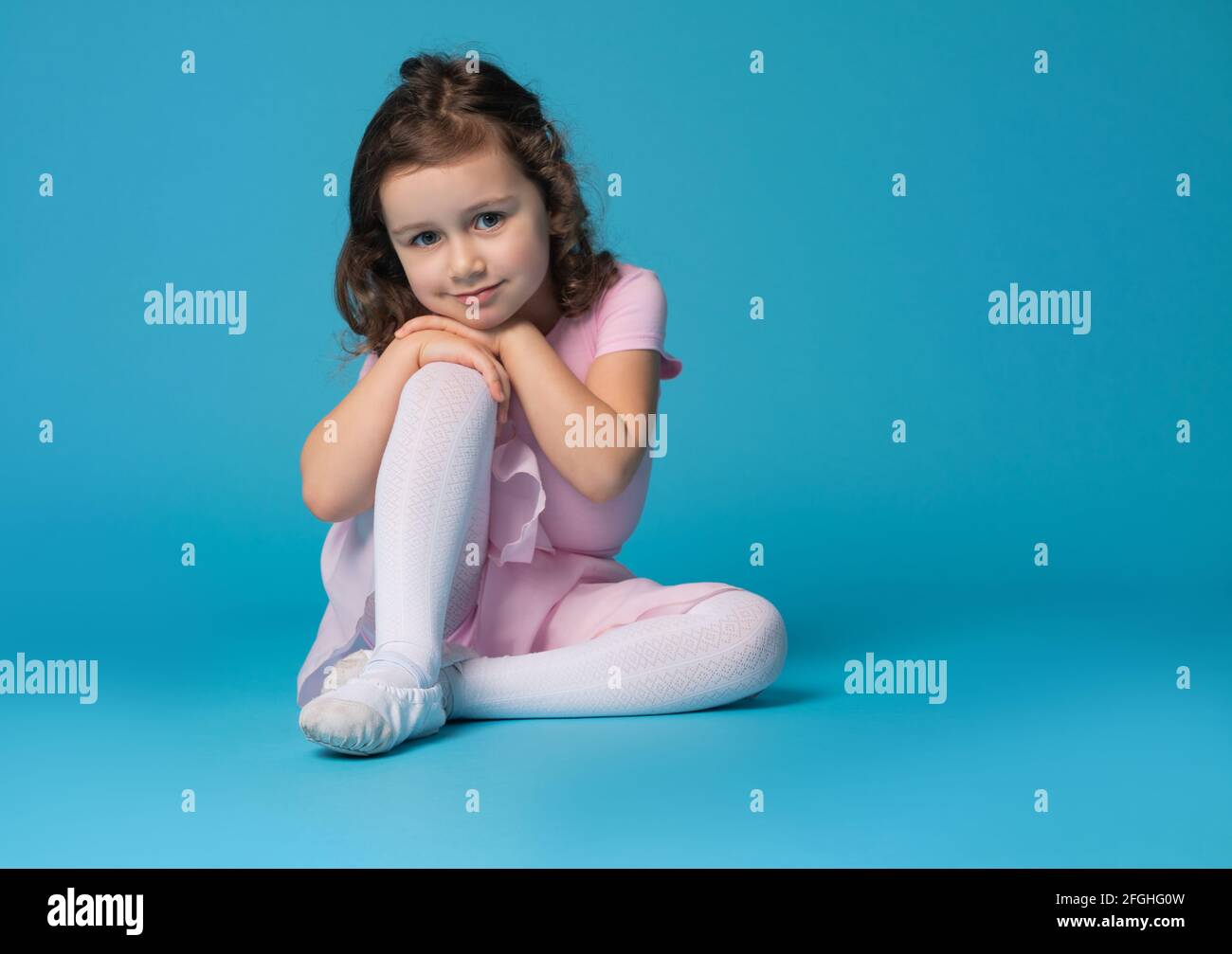 Adorable preschool girl child, ballet dancer, posing to the camera, sitting over blue background with copy space Stock Photo