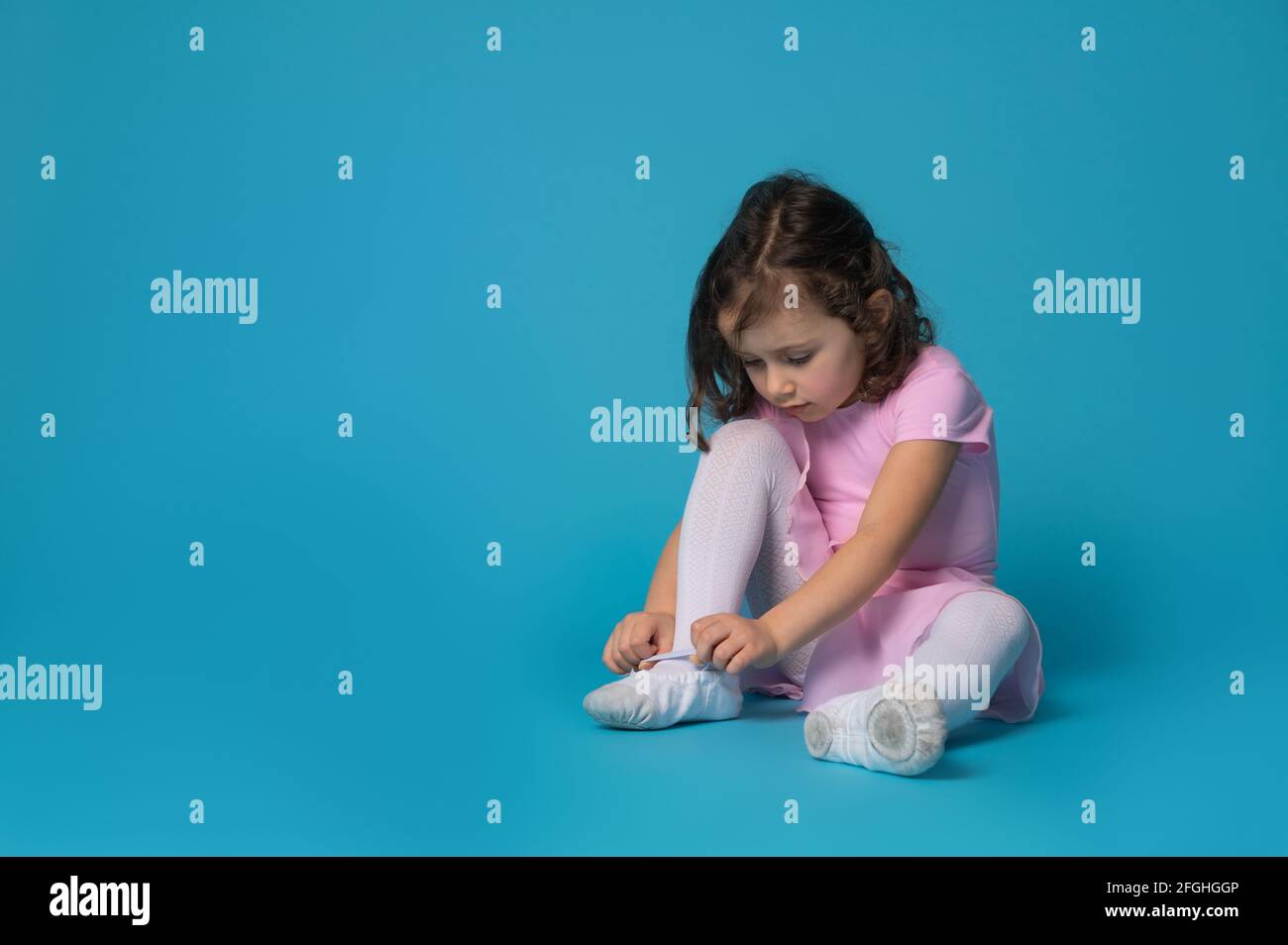 Cute ballerina focused on tying shoelaces on ballet shoes sitting on blue background with copy space Stock Photo