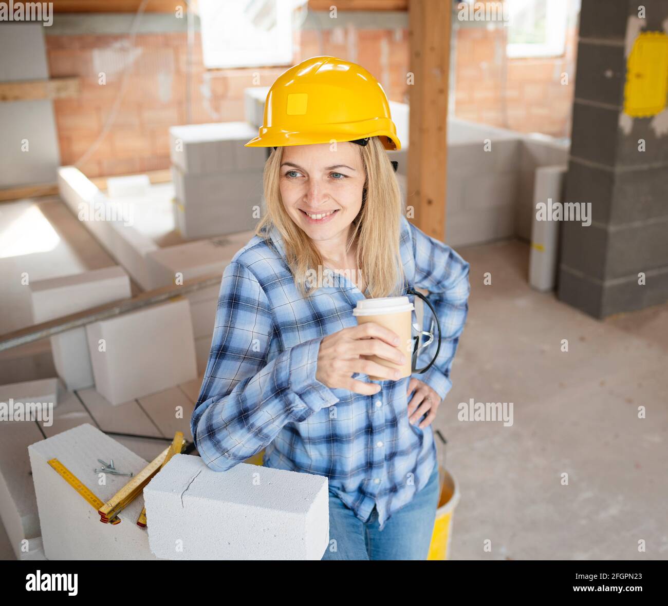 pretty young worker woman with yellow safety helmet takes coffee break on construction site indoor and is happy Stock Photo