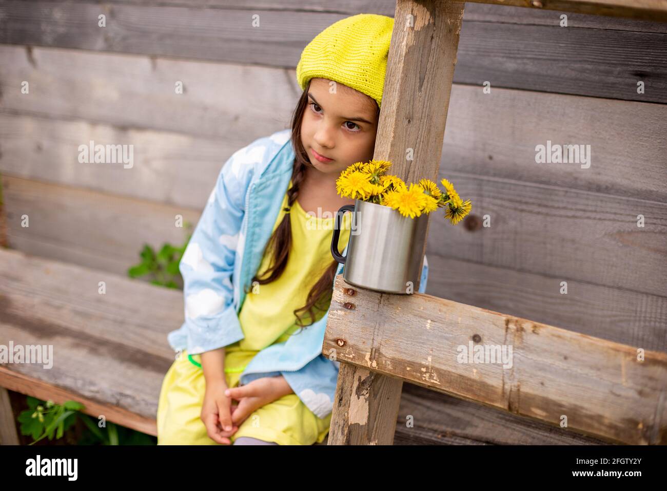 Portrait of a sad adorable little girl in a yellow beret, blue raincoat with bouquet dandelions Stock Photo