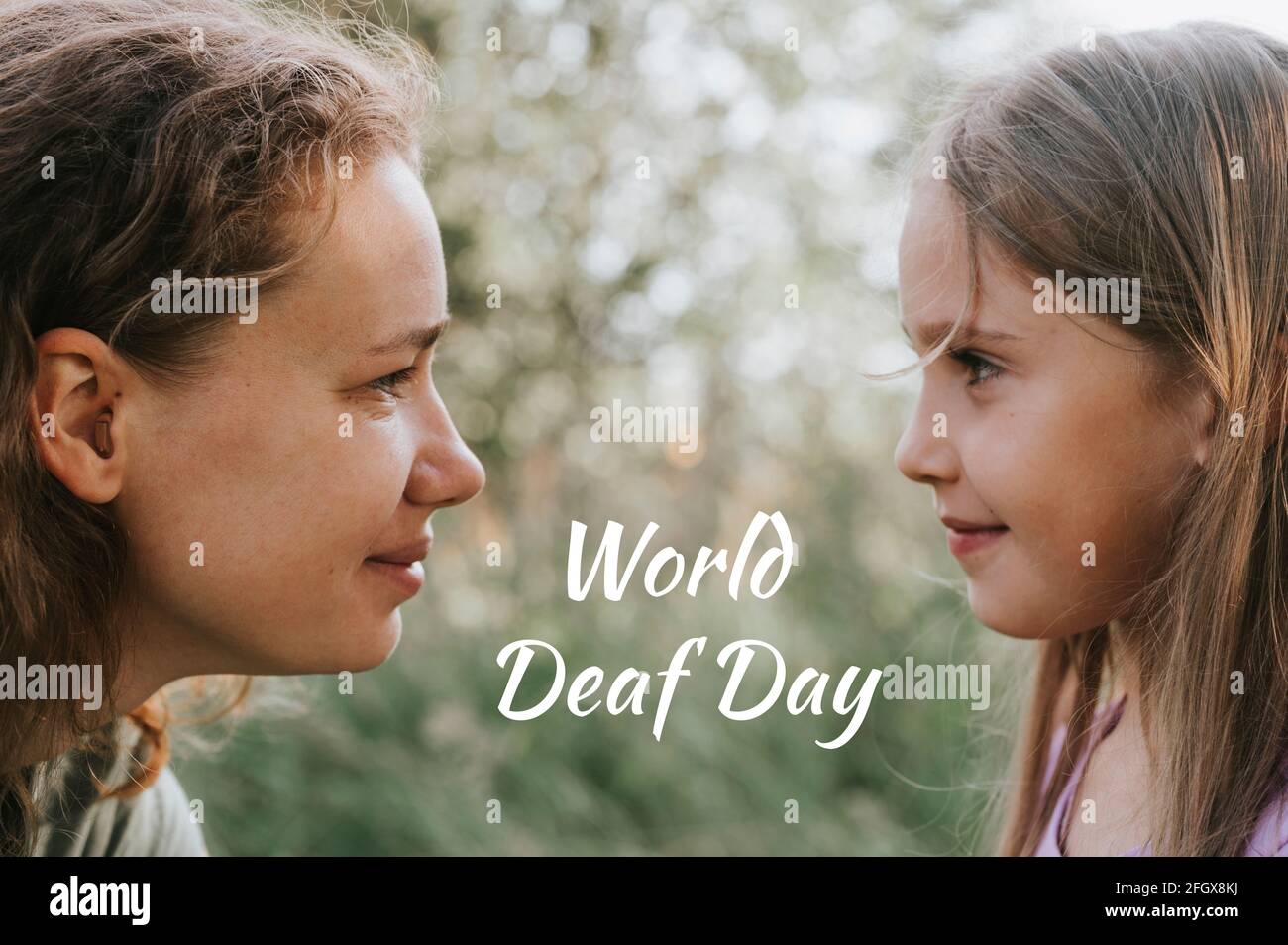 a mother with a small intra channel hearing aid communicates with her little daughter in nature outdoor. text 'World Deaf Day' OFL font Stock Photo