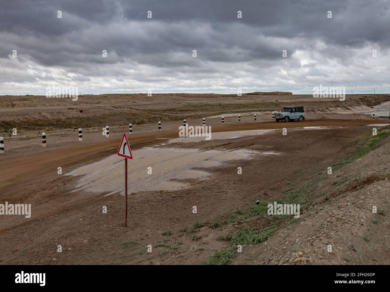 Ground road in desert after rain.Small puddle of water.Soviet military off-road car.Sign Right turn.Shardara river right.Grey storm cloudy sky. Stock Photo