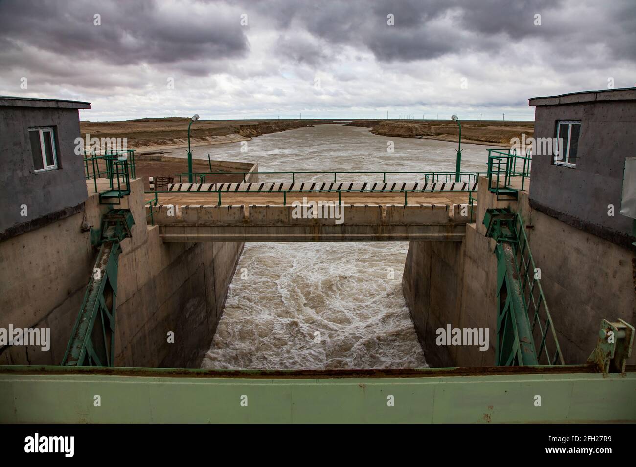 Aklak dam controls Shardara river's water level. Green metal shutter on front and bridge.Turbulent river water, grey storm clouds. Stock Photo