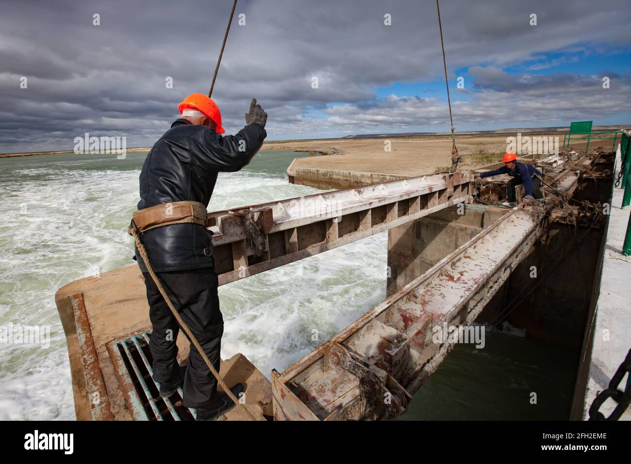 Kok-aral,Kazakhstan:Small Aral Sea dam.Workers in orange hardhat, safety belts.Lifts water shutter.Left worker show hand signal to crane operator. Stock Photo