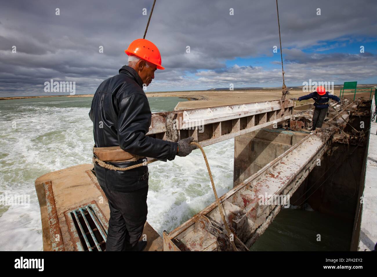 Kok-aral,Kazakhstan:Small Aral Sea Kok-aral dam.Two workers in orange helmets.Lifts water shutter weighting agent (steel girder) by crane. Stock Photo