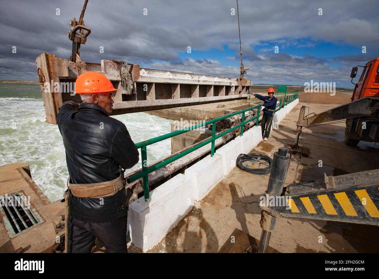 Kok-aral,Kazakhstan:Small Aral Sea dam.Two workers in orange helmets.Lifts water shutter weighting agent (steel girder) by mobile crane (right) Stock Photo