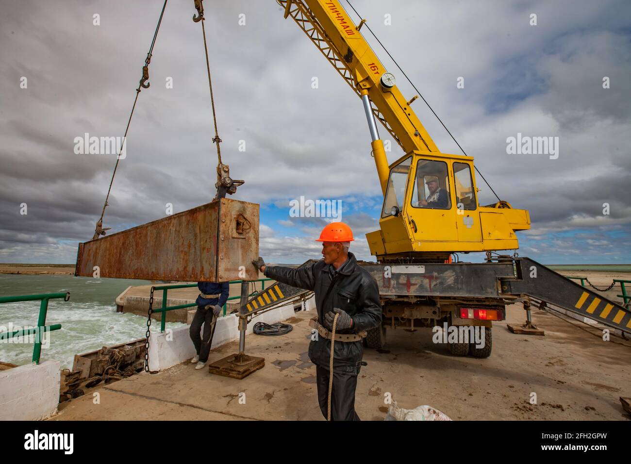 Kok-aral,Kazakhstan:Small Aral Sea Kok-aral dam.Lifting water shutter weighting agent(girder).Yellow mobile crane.Adult Asian worker in orange hardhat Stock Photo