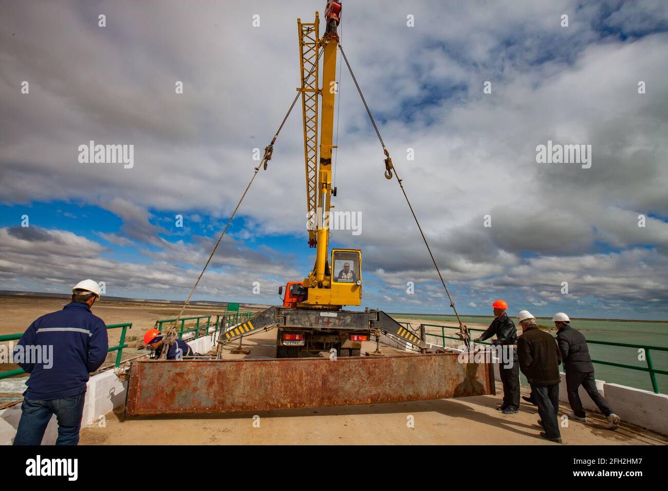 Kok-aral, Kazakhstan: Small Aral Sea Kok-aral dam. Workers lifting water shutter weighting agent. Yellow mobile crane, steel rope. Stock Photo