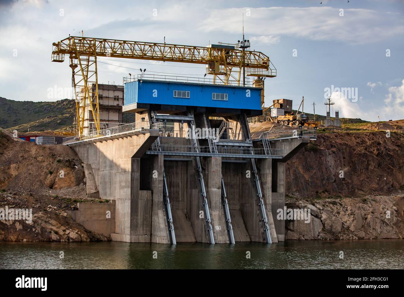 View on Sharyn river's dam. Artificial lake and dam outlets.Blue machine house, yellow gantry crane.Blue sky with clouds background.Almaty region,KZ. Stock Photo