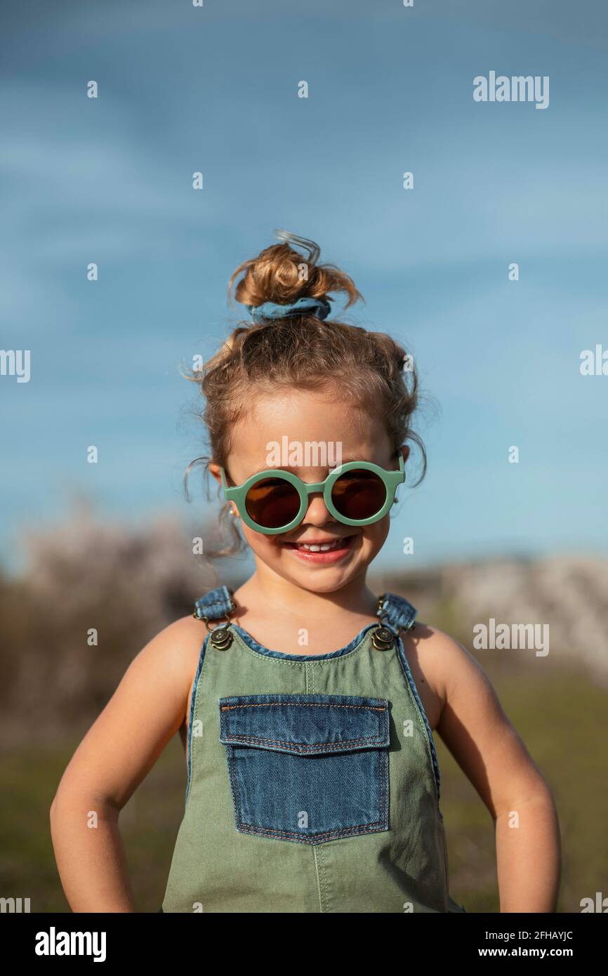 Content little girl in overalls and sunglasses standing in meadow and  enjoying summer on sunny day in countryside Stock Photo - Alamy