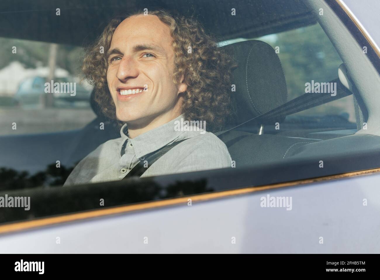 Through window view of smiling young stylish haired male passenger sitting on backseat of modern automobile and fastened with safety belt enjoying tri Stock Photo