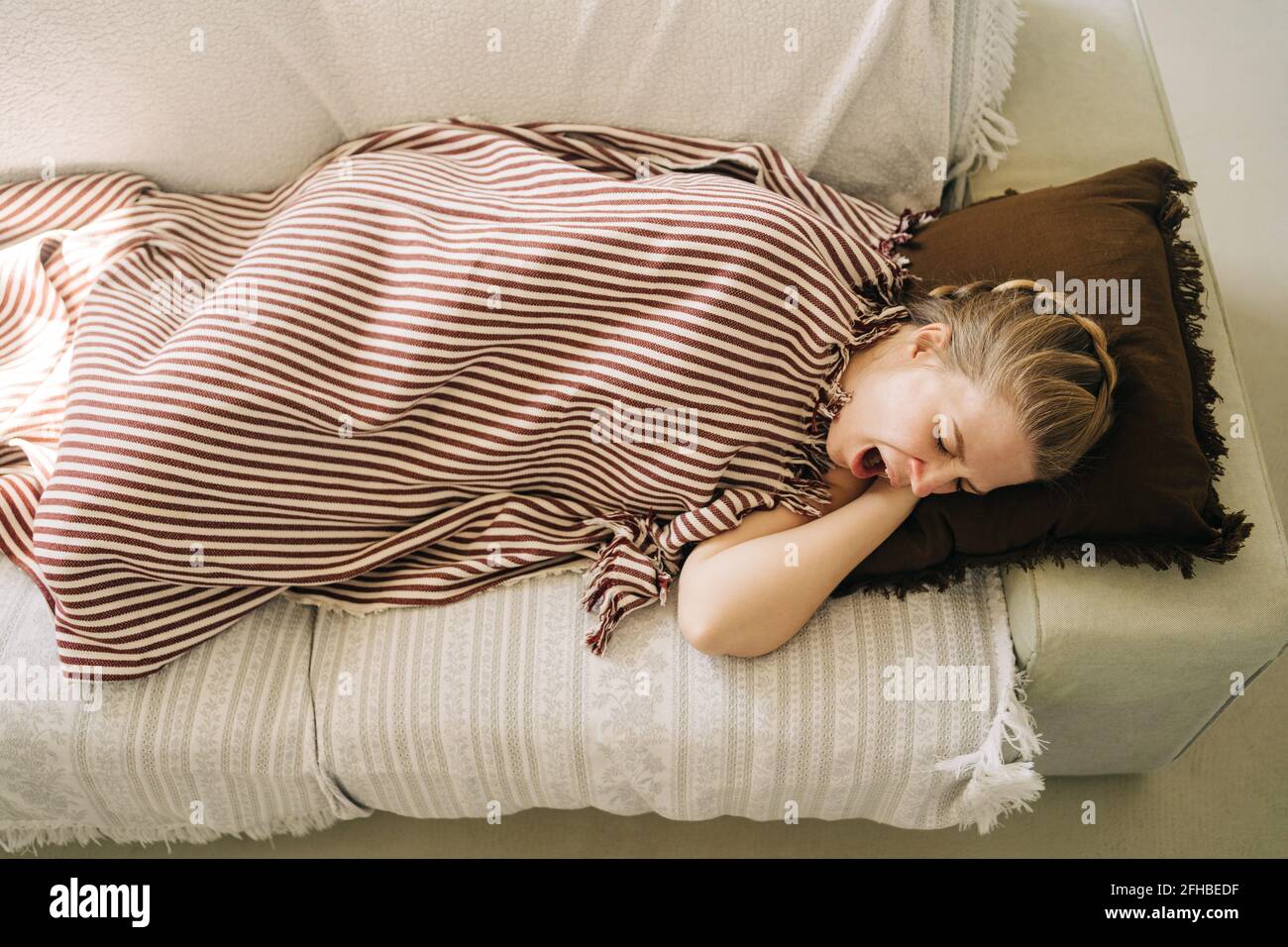 From above of young sleepy female yawning while lying under plaid on soft sofa at home Stock Photo