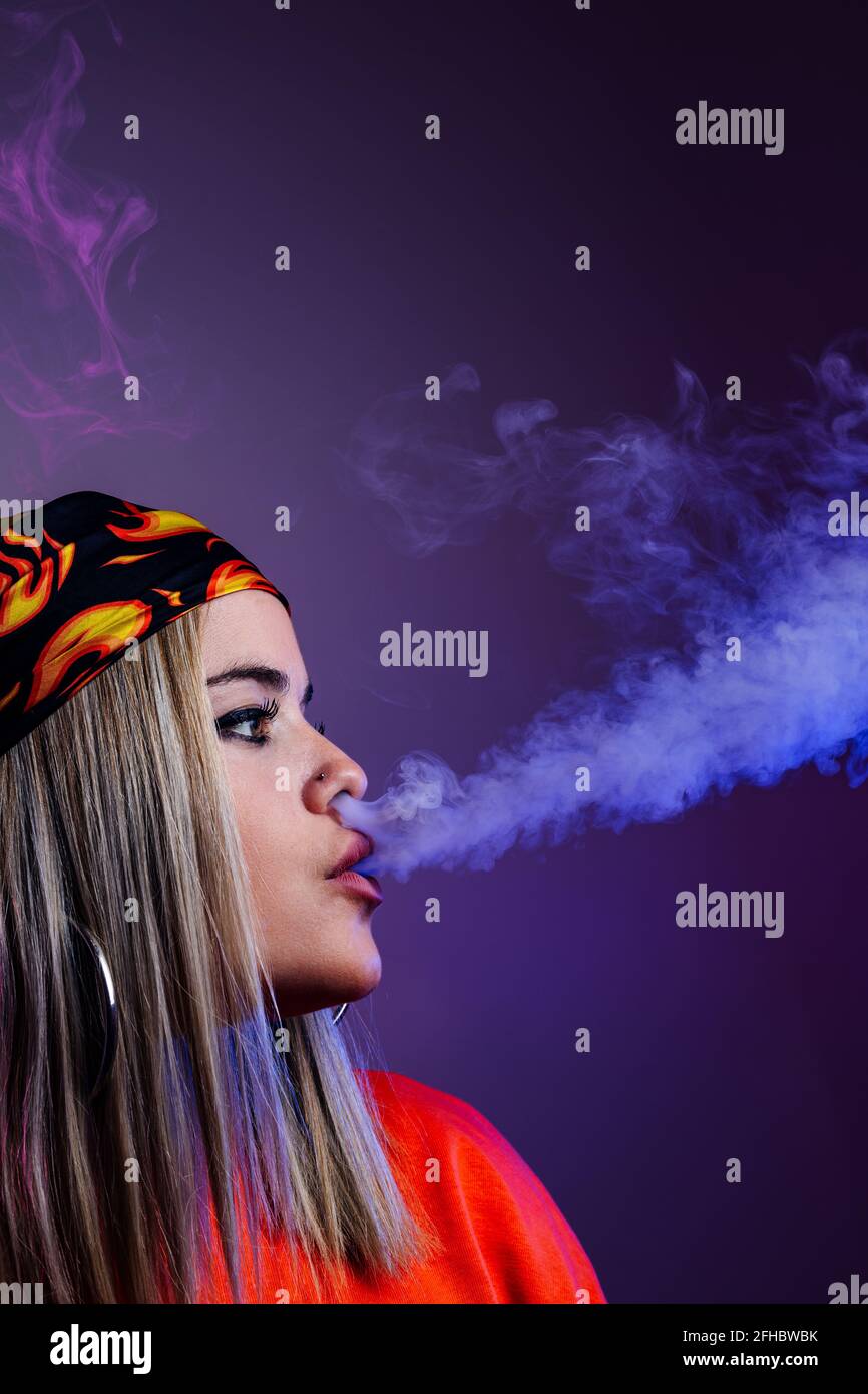 Side view of cool female in street style outfit smoking e cigarette and exhaling smoke through nose on purple background in studio with pink neon illu Stock Photo