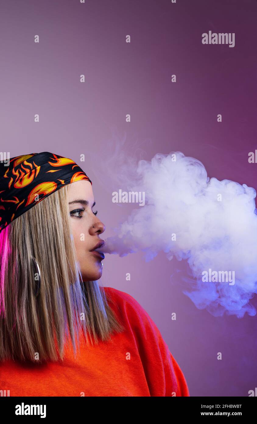 Side view of cool female in street style outfit smoking e cigarette and exhaling smoke through nose on purple background in studio with pink neon illu Stock Photo